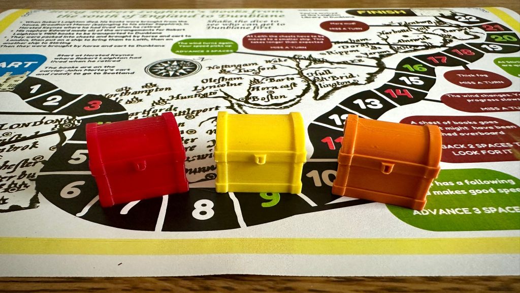 3 colourful book chest game pieces set agains a board game depicting the journey of books to the Leighton Library.